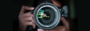 darkly lit image of a camera lens pointing directly at the viewer, with hands in shot as a header image for the blog post about how to plan a photography shoot